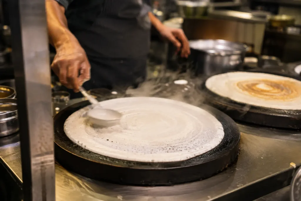 Chef preparing a dosa on a hot griddle inside a fast-service South Indian restaurant kitchen"